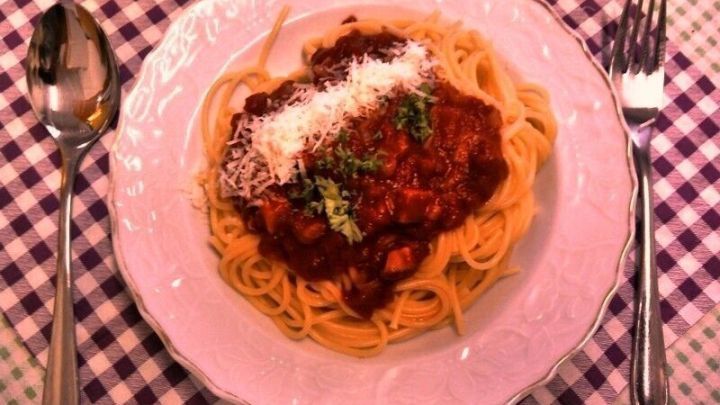 Ein Teller Spaghetti mit Tomatenso&szlig;e und Schinken ist auf einem karierten Tischdecken zu sehen, garniert mit Parmesan.