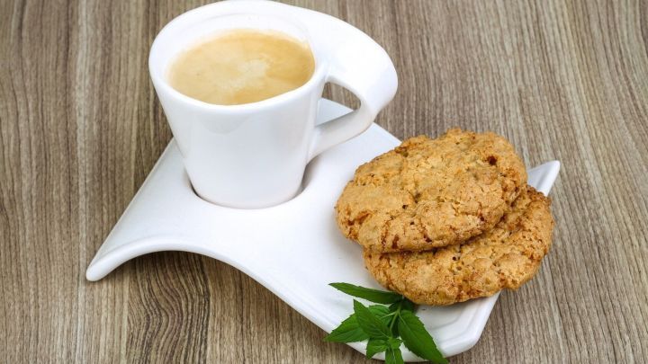 Eine wei&szlig;e Espressotasse steht mit zwei Keksen auf einem modernen Teller auf einem Holztisch. Daneben liegen frische Minzbl&auml;tter.
