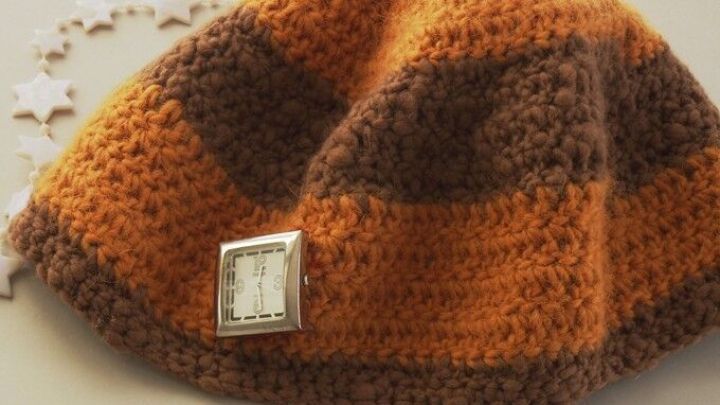 Eine geh&auml;kelte M&uuml;tze in orange und braun liegt auf einer flachen Oberfl&auml;che und hat eine Uhr an der Seite angebracht.