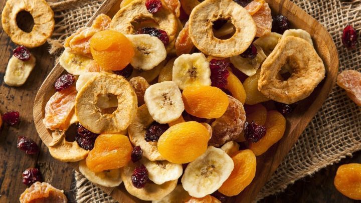 Getrocknete Fr&uuml;chte liegen auf einem Holzbrett in einer rustikalen Umgebung, darunter Bananen, Aprikosen und Apfelscheiben.