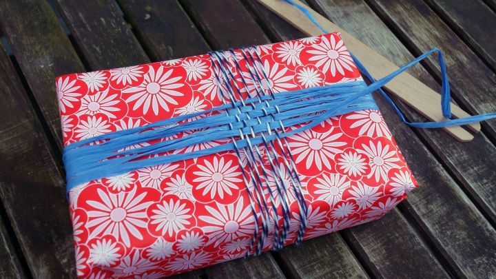 Ein dekorativ verpacktes Geschenk mit rotem Blumenmuster wird mit gewebtem Geschenkband in Blau auf einer Holztischplatte pr&auml;sentiert.