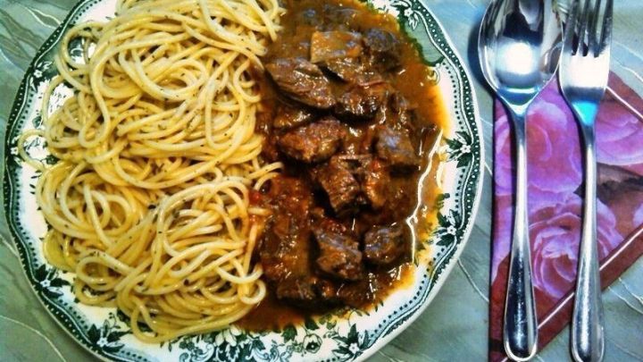 Ein Teller mit Rindergulasch und Spaghetti liegt auf einem Tisch, umgeben von Besteck und einem dekorativen Untersetzer.