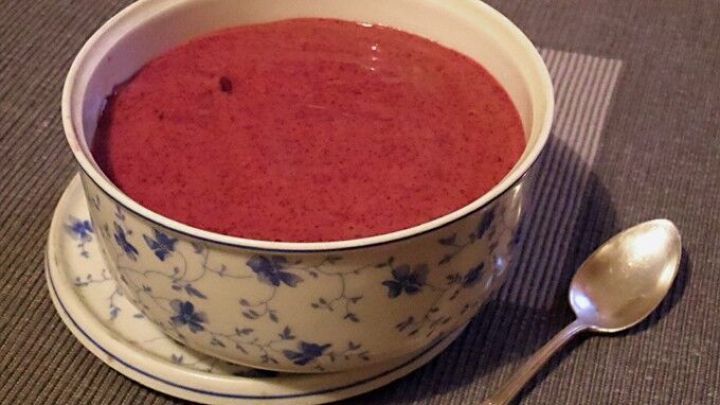 Eine Sch&uuml;ssel mit einer roten Nachspeise steht auf einem gedeckten Tisch, daneben liegt ein L&ouml;ffel auf der Tischdecke.