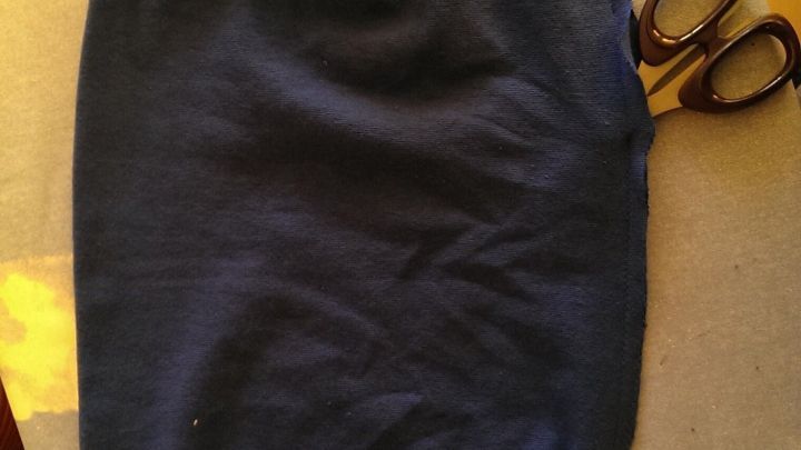 Dunkelblauer Stoffbeanie liegt auf Tisch, daneben eine Stoffschere. Hintergrund zeigt Holzboden und gr&uuml;nen Teppich.