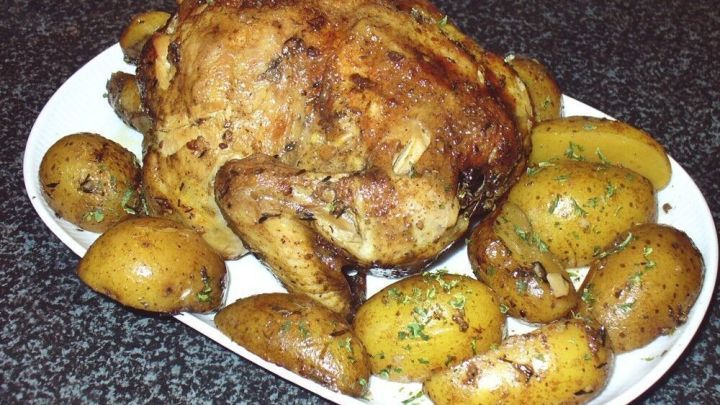 Ein goldenes H&auml;hnchen liegt auf einem wei&szlig;en Teller, umgeben von gebr&auml;unten Kartoffeln, auf einer dunklen K&uuml;chenoberfl&auml;che.