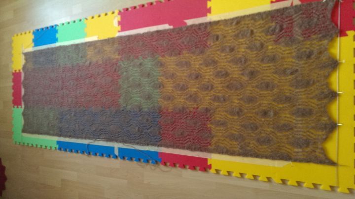 Ein Lacetuch wird auf bunten Schaumstoffplatten gespannt; dabei zeigt sich ein detailliertes Muster in einem Raum.