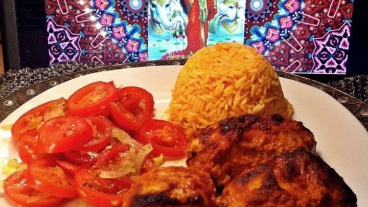 Auf einem Teller liegen geschmorte H&auml;hnchenschenkel in Joghurtso&szlig;e, umgeben von frisch geschnittenen Tomaten und Reis, vor einem bunten, indischen Hintergrund.