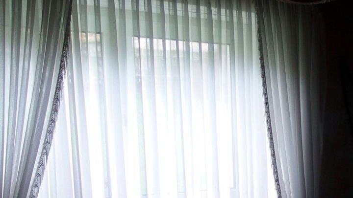 Wei&szlig;e Gardinen h&auml;ngen vor einem Fenster, das Tageslicht str&ouml;mt hindurch und beleuchtet den Raum dahinter.