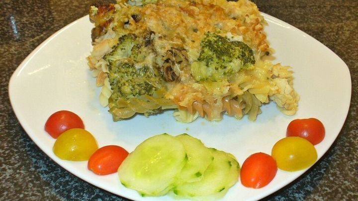 Ein Nudel-Brokkoli-Blumenkohlauflauf mit Hack liegt auf einem wei&szlig;en Teller, umgeben von frischem Gem&uuml;se als garnierte Dekoration.