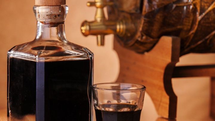 Eine Essigflasche steht neben einem Glas und einem Holzfass mit Hahn in einer warm beleuchteten Umgebung.