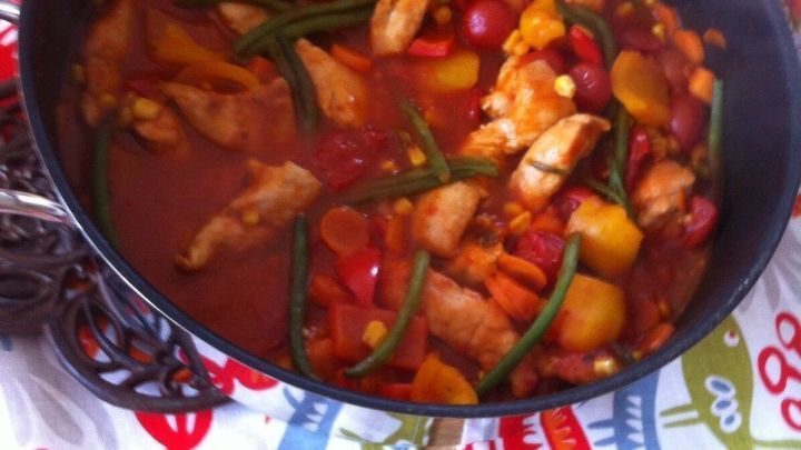 Ein bunter H&auml;hnchentopf mit Nudeln k&ouml;chelt in einer Pfanne, umgeben von einer farbenfrohen Unterlage auf einem Tisch.