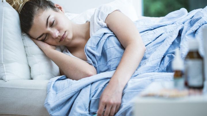 Eine erk&auml;ltete Frau liegt ersch&ouml;pft auf einer Couch, w&auml;hrend Luft in den Raum str&ouml;mt. Medikamente stehen in der N&auml;he.