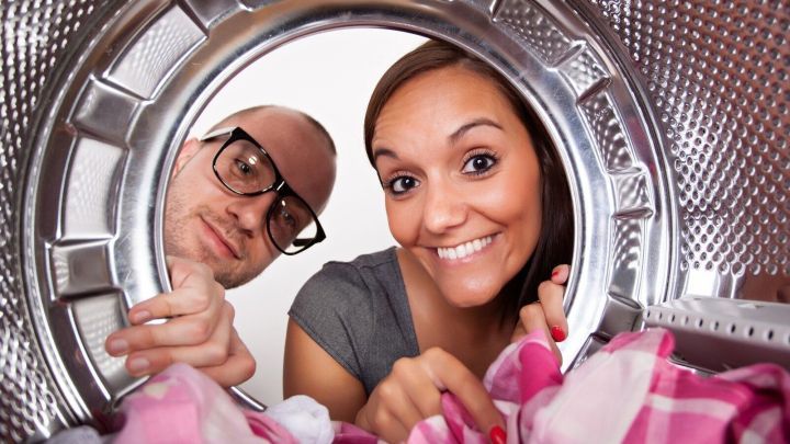 Ein Paar schaut fr&ouml;hlich durch die &Ouml;ffnung eines W&auml;schetrockners, w&auml;hrend sie farbige W&auml;sche entnehmen.