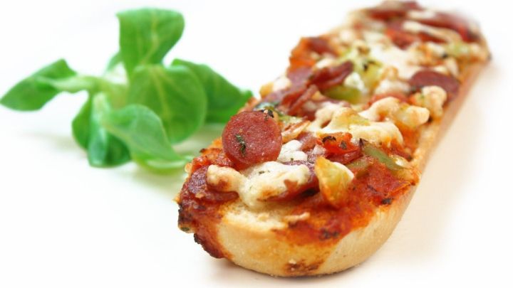 Ein knuspriges Pizzabaguette liegt auf einem wei&szlig;en Teller, auf der Seite frische Kr&auml;uter, umgeben von einem hellen Hintergrund.