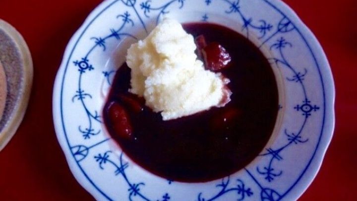 Eine Schale Fliederbeersuppe mit einer Portion heller Schlagsahne, auf einem blauen, verzierten Teller, vor rotem Hintergrund.