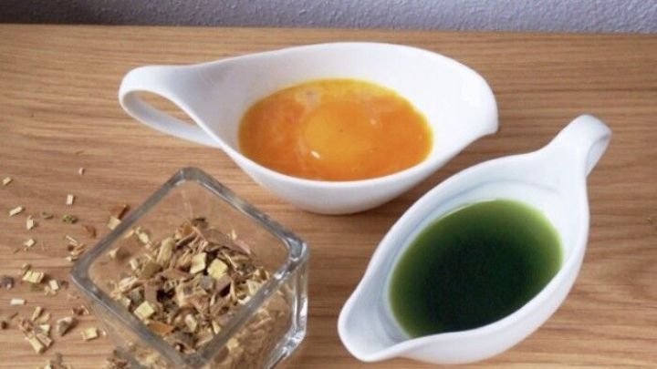 Zwei Schalen mit Haarpflege-Mischungen, eine orange und eine gr&uuml;n, stehen neben einer Glasbox mit Kr&auml;utern auf einem Holztisch.