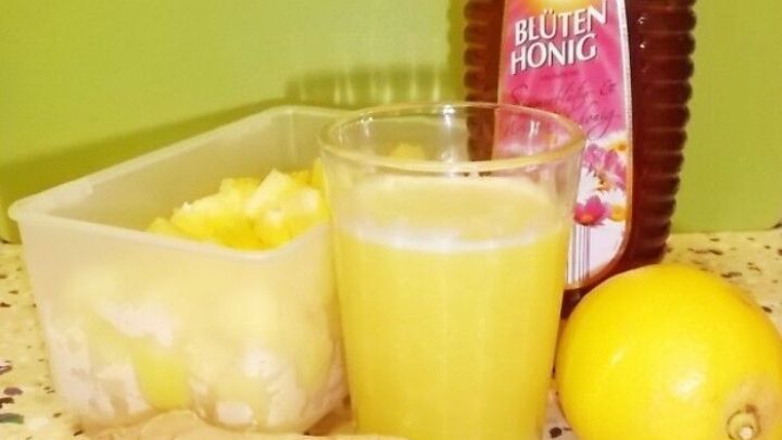 Ein Glas Ananas Hustensaft steht vor frischen Ananasst&uuml;cken, Zitrone und Honig auf einer bunten K&uuml;chenoberfl&auml;che.