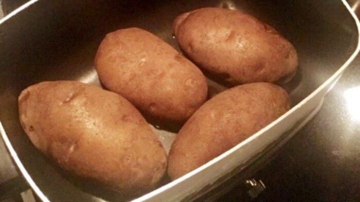 Eine wei&szlig;e Sch&uuml;ssel enth&auml;lt vier ungesch&auml;lte Kartoffeln, die auf einem Herd in einer dunklen K&uuml;che platziert sind.