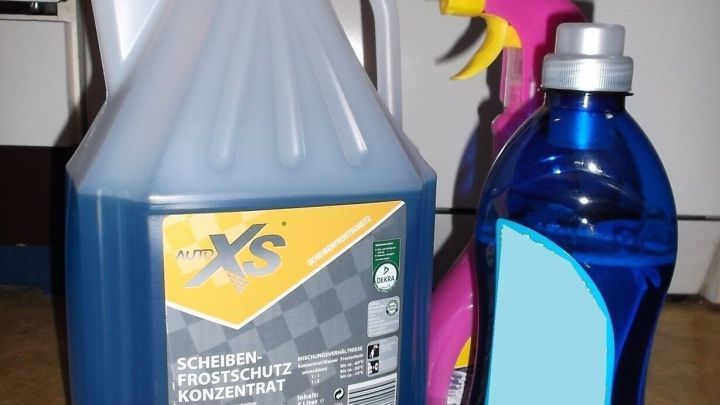 Ein blauer Kanister f&uuml;r Wasser und zwei Sprayflaschen stehen auf einem Tisch in einer Garage oder Werkstatt, bereit f&uuml;r die Scheibenwaschanlage.