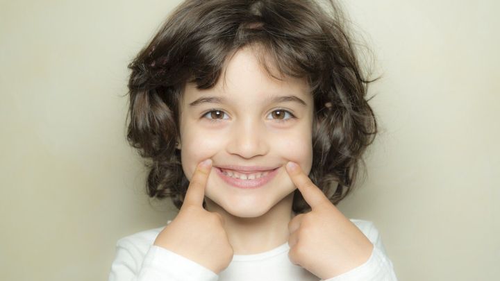 Ein lachendes Kind mit lockigem Haar zeigt seine Z&auml;hne, was auf Kreidez&auml;hne hinweisen k&ouml;nnte, vor neutralem Hintergrund.