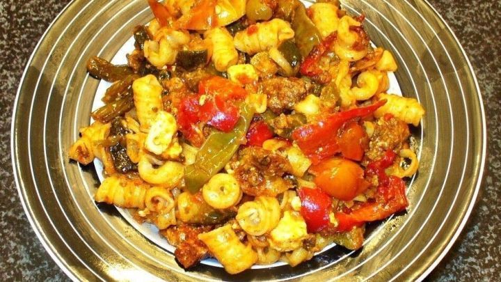 Eine bunte Nudel-Hack-Gem&uuml;sepfanne mit Fetak&auml;se auf einem silbernen Teller, arrangiert auf einer dunklen Arbeitsfl&auml;che.