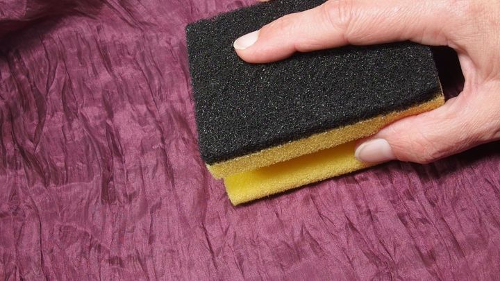 Ein Hand mit einem K&uuml;chenschwamm entfernt Fusseln von einem lila, strukturierten Stoff in einem h&auml;uslichen Umfeld.