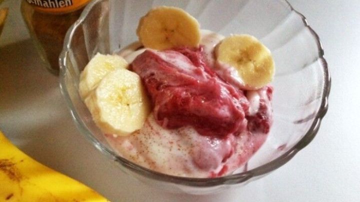 In einer Glassch&uuml;ssel liegt cremiger Naturjoghurt mit Scheiben einer Banane und roter Marmelade, umgeben von einer Banane und Zimt.