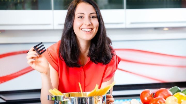 Eine l&auml;chelnde Frau in einem modernen K&uuml;chensetup h&auml;lt einen Kochl&ouml;ffel &uuml;ber einem Topf, umgeben von frischen Zutaten.