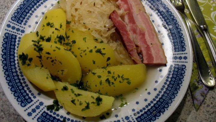 Auf einem Teller liegen gut zubereitete Salzkartoffeln, serviert mit Sauerkraut und Schweinefleisch, in einer gem&uuml;tlichen K&uuml;chenszene.