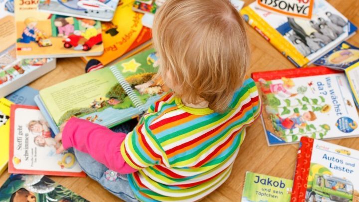 Ein Kind sitzt auf dem Boden und bl&auml;ttert in einem Bilderbuch, umgeben von vielen bunten Bilderb&uuml;chern zum Vorlesen.