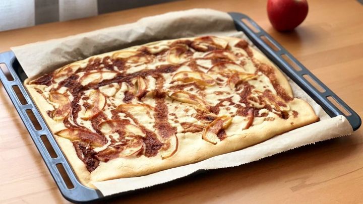 Apfelpfannkuchen auf Blech, gebacken mit Zimt und Apfelscheiben, steht auf einem Tisch neben einem roten Apfel.