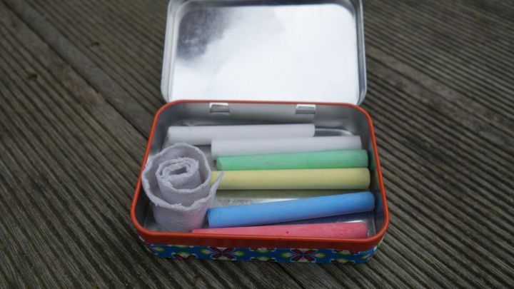 Eine Blechdose ist ge&ouml;ffnet, enth&auml;lt verschiedene Malutensilien und liegt auf einem Holztisch im Freien.