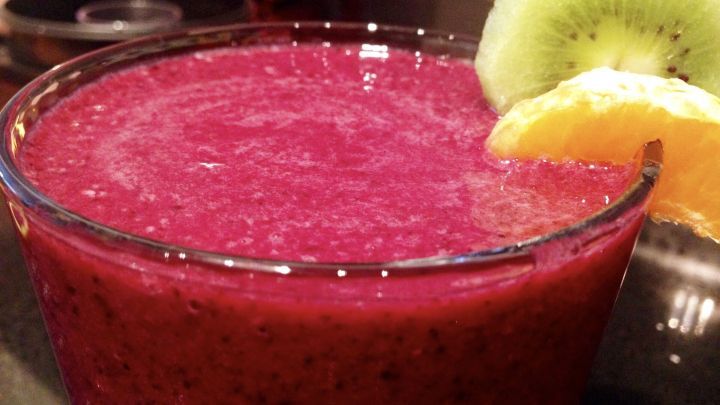 Ein leuchtend lila Blaubeer-Bananen-Smoothie steht in einem Glas und wird von einer Kiwi und einer Orangenscheibe dekoriert.