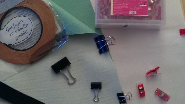 Eine schwarze Gurtbandrolle liegt auf einem Tisch, umgeben von verschiedenen B&uuml;roklammern und Wachstuchst&uuml;cken zum Verarbeiten.