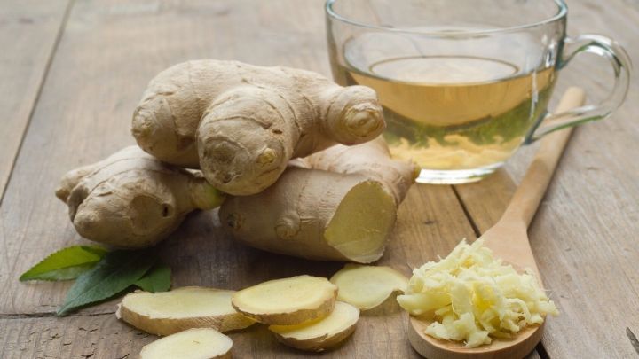 Frische Ingwerknollen liegen neben einer Tasse Ingwertee und einem L&ouml;ffel mit geriebenem Ingwer auf einem Holztisch.