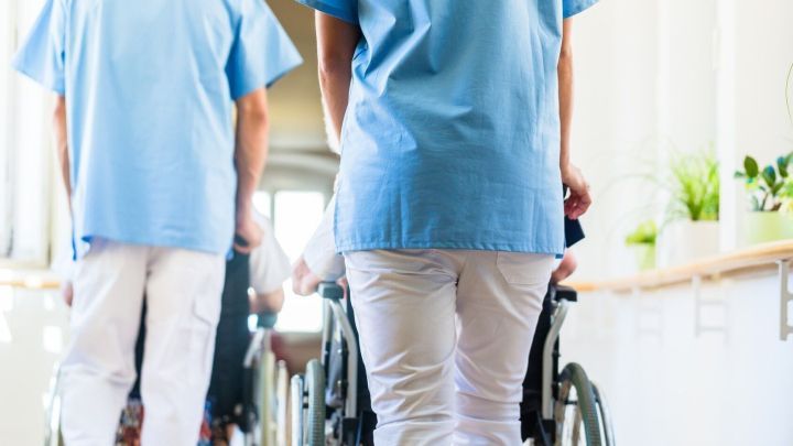 Pflegekr&auml;fte in blauen Uniformen schieben Rollst&uuml;hle durch einen hellen Flur in einem Pflegeheim, um Bewohner zu unterst&uuml;tzen.