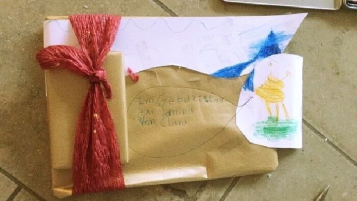 Ein mit buntem Papier verpacktes Paket liegt auf einem Tisch, dekoriert mit Kinderzeichnungen und orangefarbener Schnur.