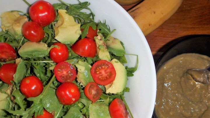Ein frischer Avocado-Tomate-Rucola-Salat liegt in einer Sch&uuml;ssel. Daneben ist eine Banane und ein Honigdressing in einer Schale.