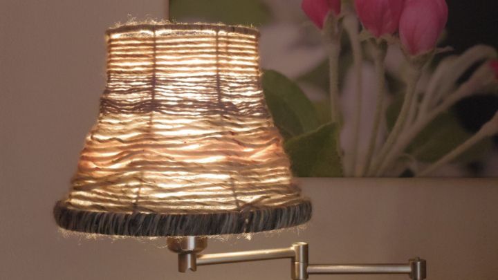 Ein Lampenschirm, mit Wollgarn umwickelt, leuchtet sanft in einem Raum mit einer Blumenfotografie im Hintergrund.