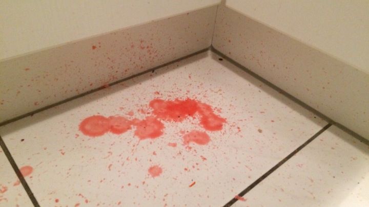 Dunkelrotes Kerzenwachs ist auf wei&szlig;en Fliesen versch&uuml;ttet, mit Tropfen und Spritzern &uuml;berall im Eckenbereich verteilt.