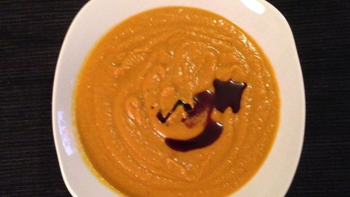 Eine sch&uuml;ssel K&uuml;rbis-Curry-Kokos-Chili-Suppe steht vor schwarzem Hintergrund. Die glatte, orangefarbene Suppe hat einen dekorativen Strich aus dunkler So&szlig;e.