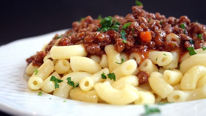 H&ouml;rnchennudeln mit Hackfleisch liegen auf einem wei&szlig;en Teller, begleitet von einer herzhaften Hackfleischsauce und frischem Gr&uuml;n.