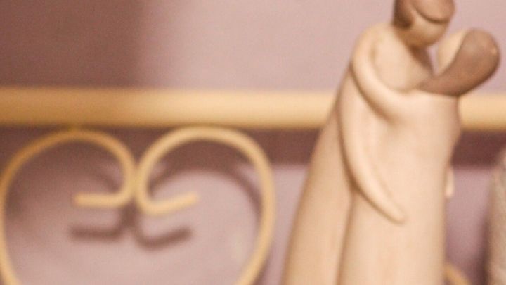 Tonfigur eines umarmenden Paares vor einem herzf&ouml;rmigen Metallgitter, unscharfer lila Hintergrund.