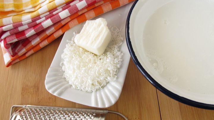 Auf einem Holztisch liegen einige Zutaten zum selbst herstellen von biologischem Waschmittel: eine Sch&uuml;ssel mit Wasser, geriebene Seife und ein St&uuml;ck Seife. Im Hintergrund sind karierte Geschirrt&uuml;cher zu sehen.