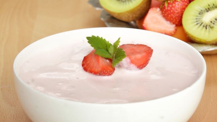 Eine Sch&uuml;ssel Joghurt mit Erdbeeren und Minze oben drauf steht auf einem Tisch, umgeben von frischem Obst.