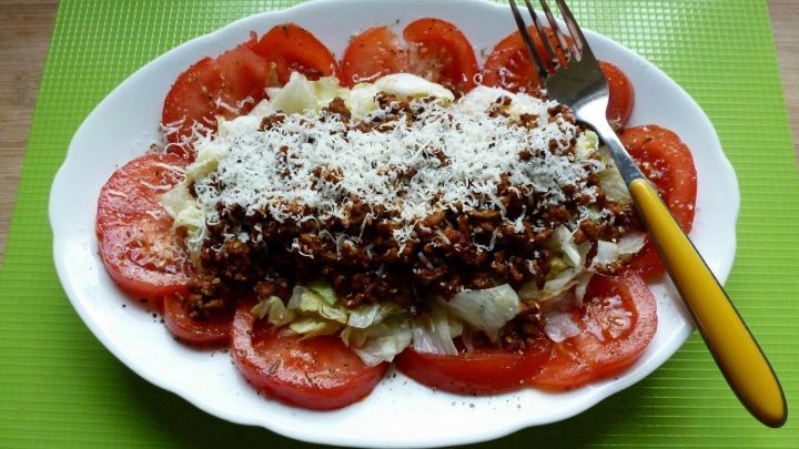 Ein Salat im Stil eines Big Mac, dekoriert mit Tomatenscheiben und K&auml;se, liegt auf einem hellgr&uuml;nen Tisch.