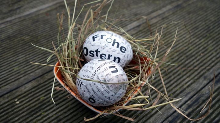 Zwei Ostereier aus Zeitungspapier liegen in einem kleinen Nest aus Heu auf einem Holztisch.
