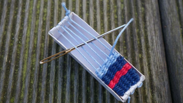 Mini-Webrahmen aus Streichholzschachtel basteln: Ein handgemachter Webrahmen mit Garn und Nadel auf einer h&ouml;lzernen Oberfl&auml;che.