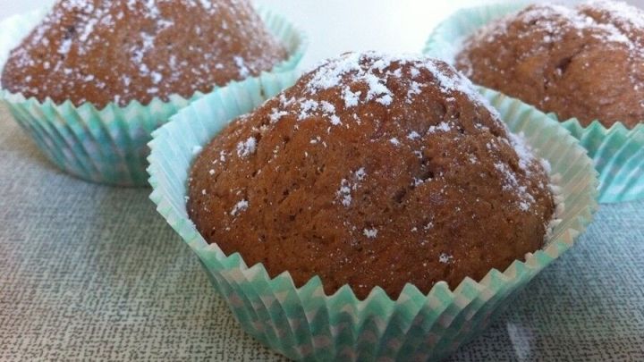 Drei Bananen-Schoko-Muffins liegen in hellblauen Papierf&ouml;rmchen auf einer neutralen Oberfl&auml;che und sind mit Puderzucker best&auml;ubt.