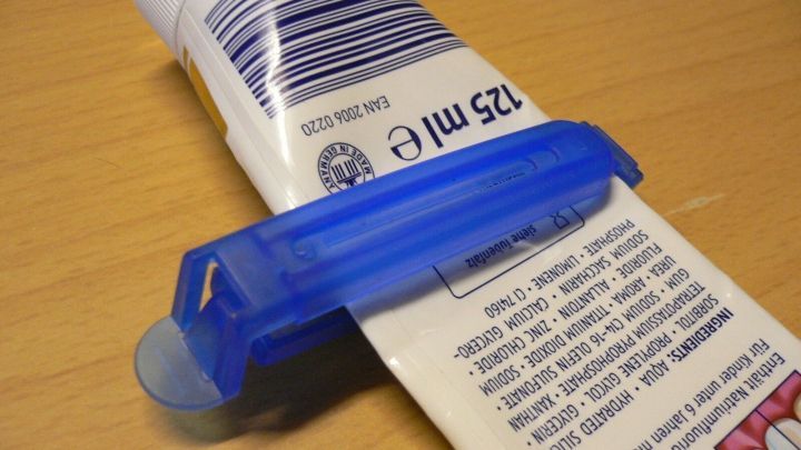 Eine blaue Kunststofftubenausdr&uuml;ckhilfe presst eine Zahnpastatube auf einem Tisch, um den Inhalt effektiv zu entleeren.